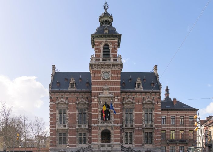Un grand bâtiment orné de briques et de pierres avec une tour d'horloge centrale et une entrée voûtée, arborant trois drapeaux au-dessus des escaliers, situé sur une vaste place sous un ciel bleu.