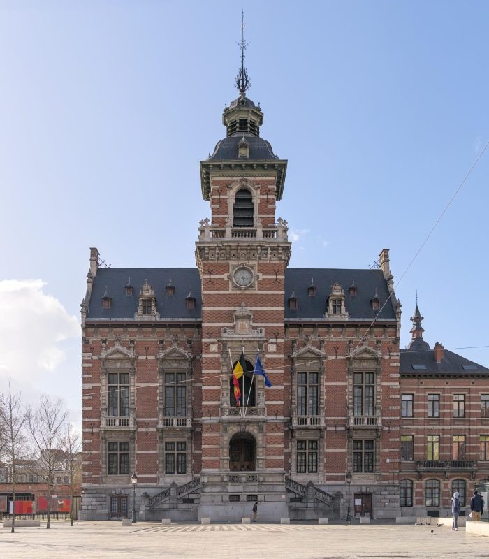 Un grand bâtiment orné de briques et de pierres avec une tour d'horloge centrale et une entrée voûtée, arborant trois drapeaux au-dessus des escaliers, situé sur une vaste place sous un ciel bleu.