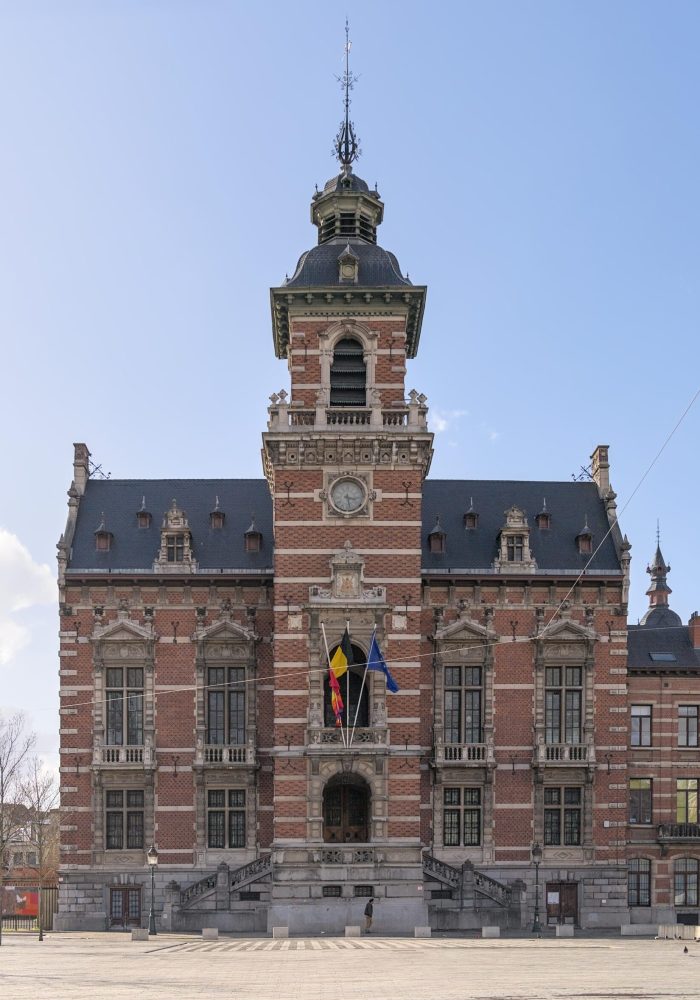 Un grand bâtiment orné de briques et de pierres avec une tour d'horloge centrale et une entrée voûtée, arborant trois drapeaux au-dessus des escaliers, situé sur une vaste place sous un ciel bleu.