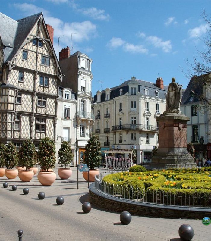 A European town square with a statue surrounded by yellow flowers, white half-timbered historic buildings, potted trees and a blue sky with scattered clouds.