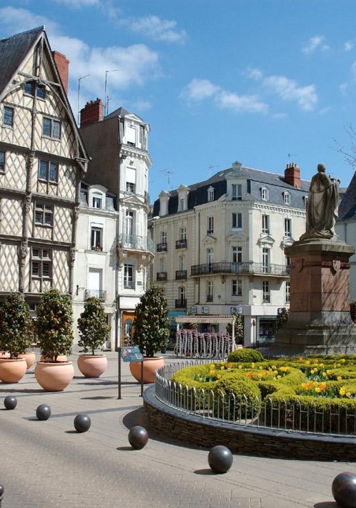 Une place de ville européenne avec une statue entourée de fleurs jaunes, des bâtiments historiques à colombages et blancs, des arbres en pot et un ciel bleu avec des nuages épars.
