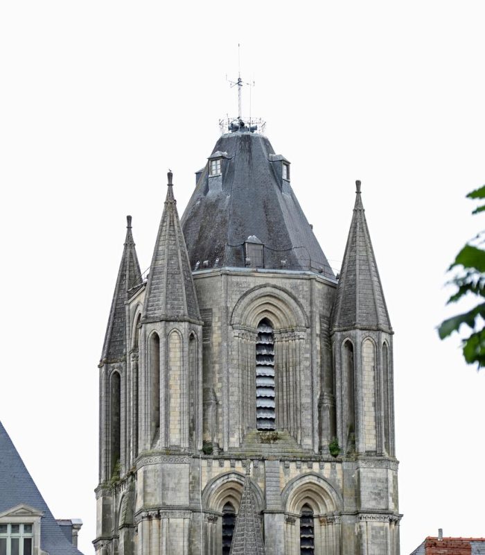 Un haut clocher en pierre avec des fenêtres arquées et quatre flèches pointues, entouré par les toits des bâtiments voisins, sur fond de ciel blanc couvert.