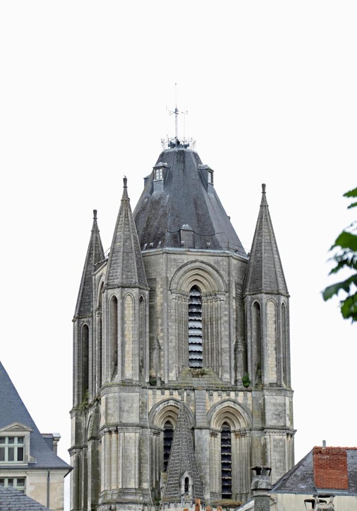 A tall stone bell tower with arched windows and four pointed spires, surrounded by the roofs of neighbouring buildings, against a backdrop of an overcast white sky.
