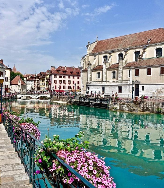 Un canal pittoresque à l'eau turquoise traverse une ville européenne bordée de bâtiments colorés, de fleurs, de piétons et d'une église au clocher élevé sous un ciel partiellement nuageux.