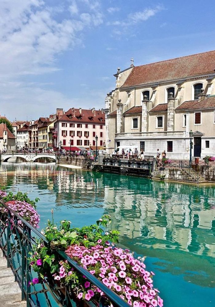 Un canal pittoresque à l'eau turquoise traverse une ville européenne bordée de bâtiments colorés, de fleurs, de piétons et d'une église au clocher élevé sous un ciel partiellement nuageux.