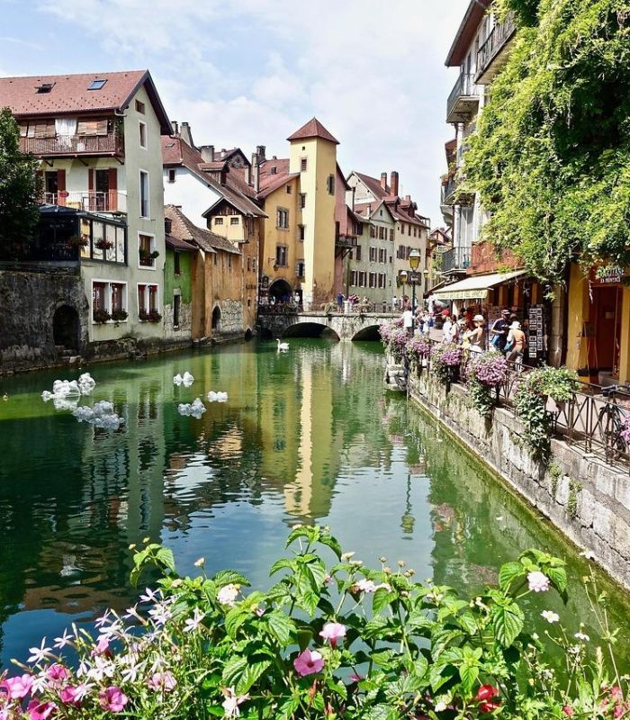 Un charmant canal bordé de bâtiments colorés, de bacs à fleurs et d'un petit pont, reflétant le paysage dans l'eau. Les gens se promènent le long de la promenade en pierre, à côté des boutiques et des cafés, lors d'une journée ensoleillée.