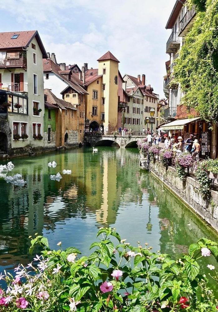 Un charmant canal bordé de bâtiments colorés, de bacs à fleurs et d'un petit pont, reflétant le paysage dans l'eau. Les gens se promènent le long de la promenade en pierre, à côté des boutiques et des cafés, lors d'une journée ensoleillée.