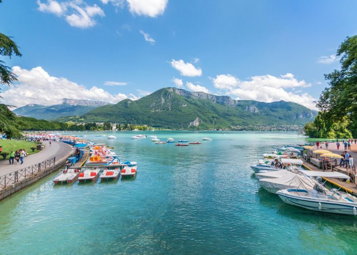 Vue panoramique d'un lac turquoise avec des bateaux amarrés le long des deux rives, des personnes marchant sur des sentiers et des montagnes verdoyantes en arrière-plan, sous un ciel partiellement nuageux.