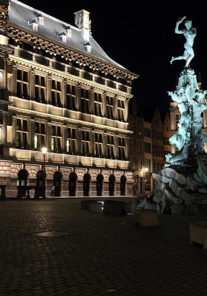 Une place de ville, la nuit, avec de grands bâtiments historiques illuminés et une statue proéminente d'un personnage au sommet d'un socle rocheux au centre. La zone est vide et pavée.