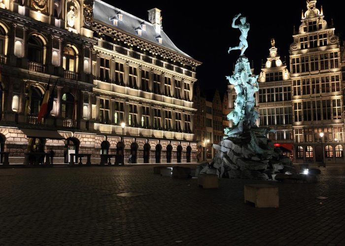 A town square, at night, with large illuminated historic buildings and a prominent statue of a figure atop a rocky plinth in the centre. The area is empty and paved.