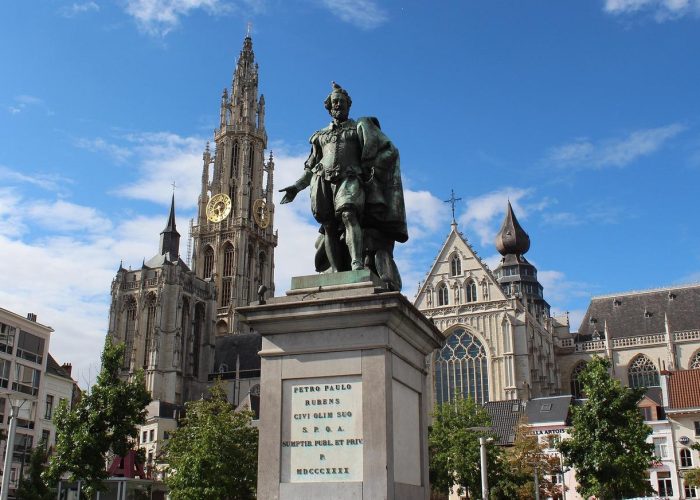Une statue en bronze se dresse sur un piédestal en pierre sur une place de la ville, entourée d'arbres. À l'arrière-plan, on aperçoit une grande cathédrale gothique avec une haute tour d'horloge et une architecture ornée sous un ciel bleu.