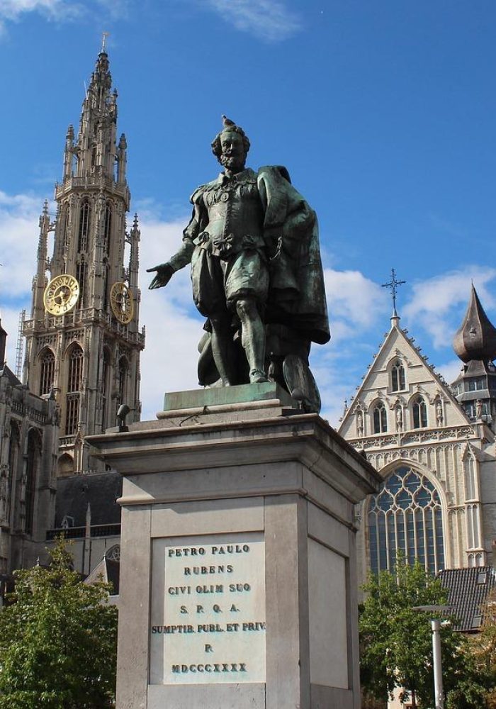 A bronze statue stands on a stone pedestal in a town square, surrounded by trees. In the background is a large Gothic cathedral with a tall clock tower and ornate architecture beneath a blue sky.