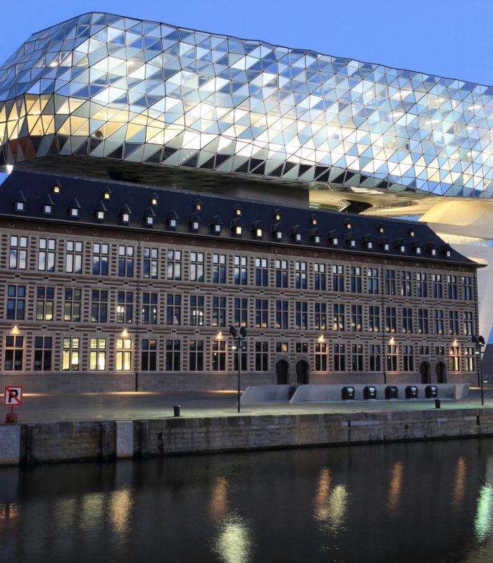 Un bâtiment historique surmonté d'une structure géométrique moderne en verre, éclairée au crépuscule et se reflétant dans l'eau d'un canal situé en face.