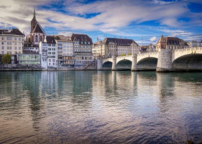 Un pont de pierre enjambe une rivière calme, avec des bâtiments historiques et une église dotée d'un haut clocher au bord de l'eau, sous un ciel partiellement nuageux.