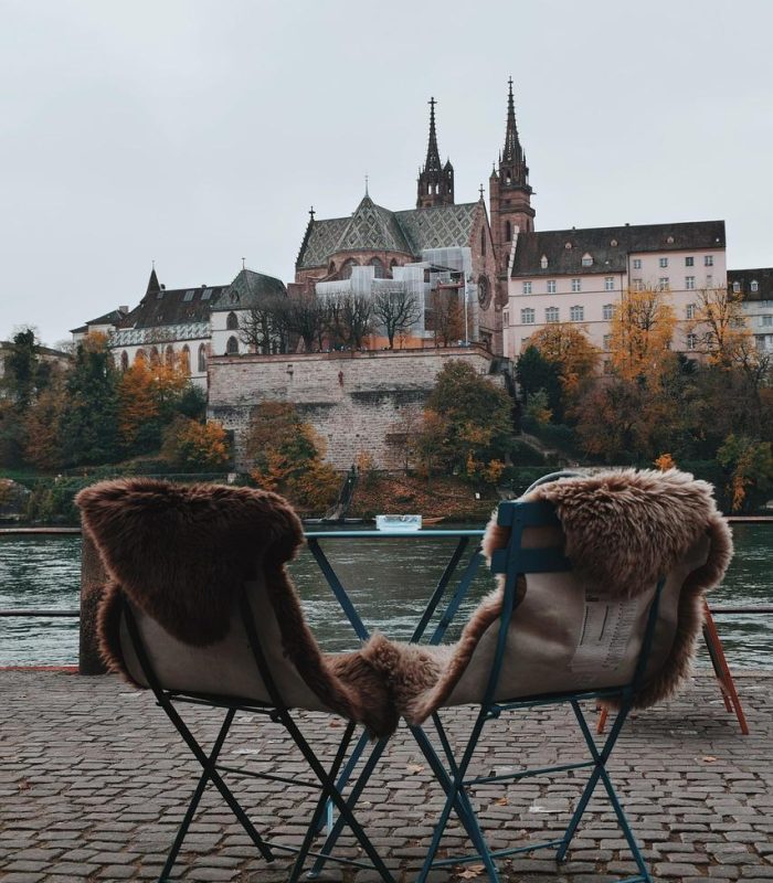 Deux chaises recouvertes de fourrure et une petite table sont assises sur un chemin pavé au bord d'une rivière, avec un vieux bâtiment de style gothique et des arbres d'automne en arrière-plan sous un ciel nuageux.