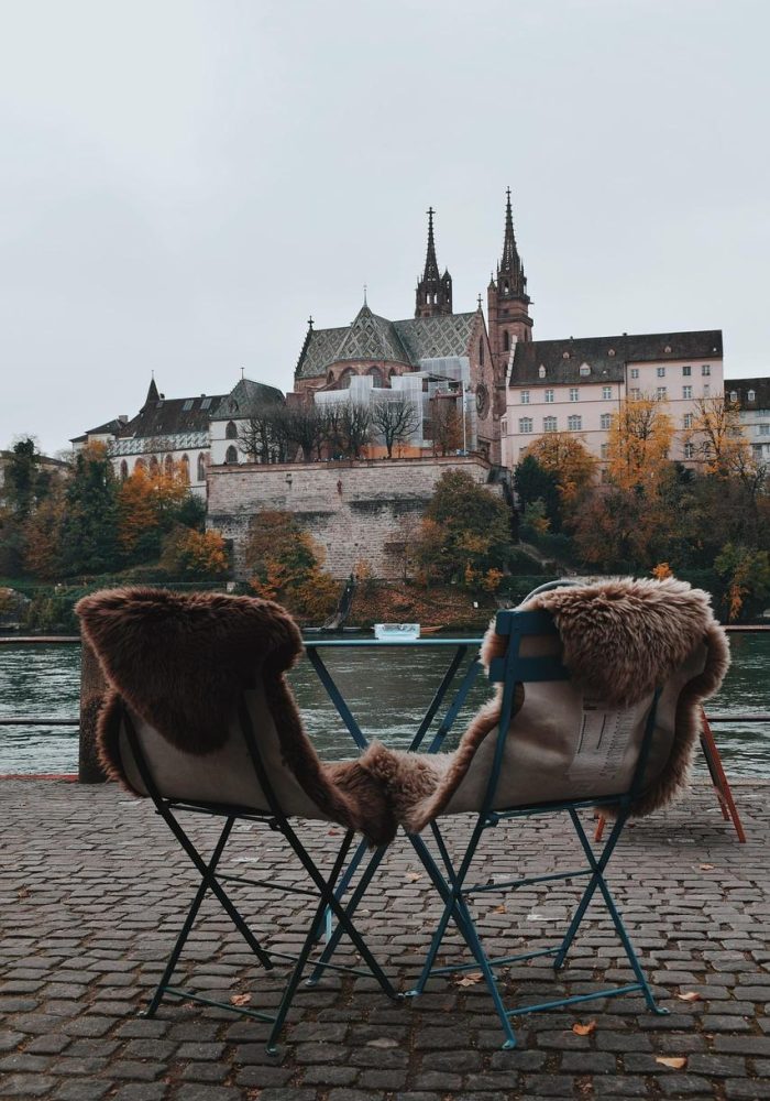 Deux chaises recouvertes de fourrure et une petite table sont assises sur un chemin pavé au bord d'une rivière, avec un vieux bâtiment de style gothique et des arbres d'automne en arrière-plan sous un ciel nuageux.