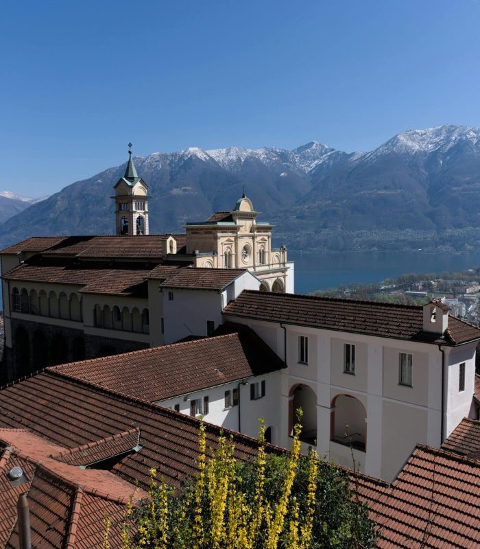 Un bâtiment historique aux toits de tuiles rouges se dresse sur une colline surplombant un grand lac, avec des montagnes enneigées et un ciel bleu clair en arrière-plan.