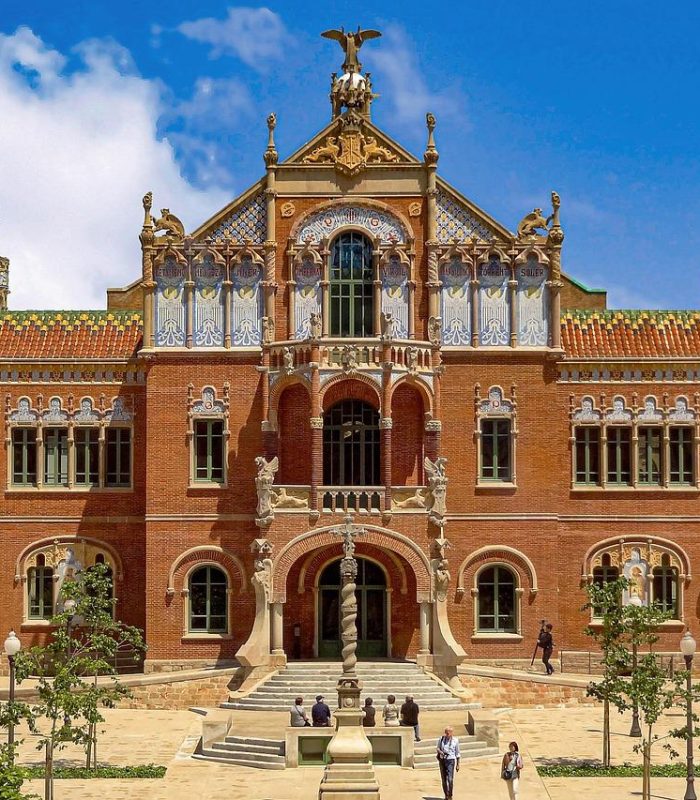Un grand bâtiment historique à l'architecture ornée, aux murs de briques rouges, aux fenêtres cintrées et aux toits en dôme colorés, sur fond de ciel bleu vif, avec des gens qui marchent et de la verdure au premier plan.