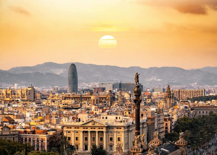 Vue panoramique de Barcelone au coucher du soleil, avec des bâtiments historiques, le monument de Christophe Colomb et des montagnes lointaines derrière lesquelles le soleil se couche, projetant une lumière chaude et dorée sur la ville.