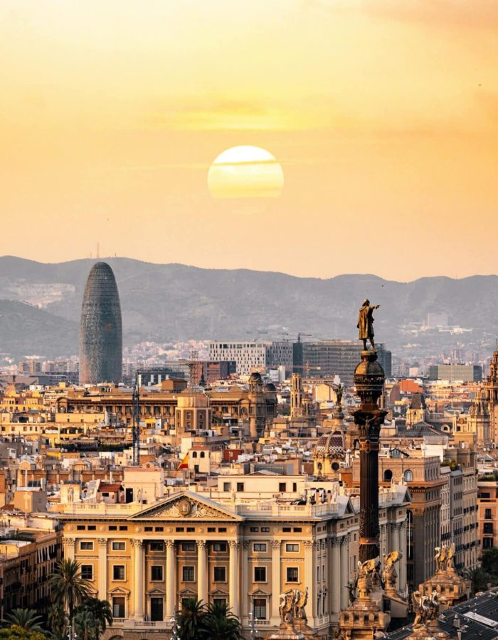 Vue panoramique de Barcelone au coucher du soleil, avec des bâtiments historiques, le monument de Christophe Colomb et des montagnes lointaines derrière lesquelles le soleil se couche, projetant une lumière chaude et dorée sur la ville.