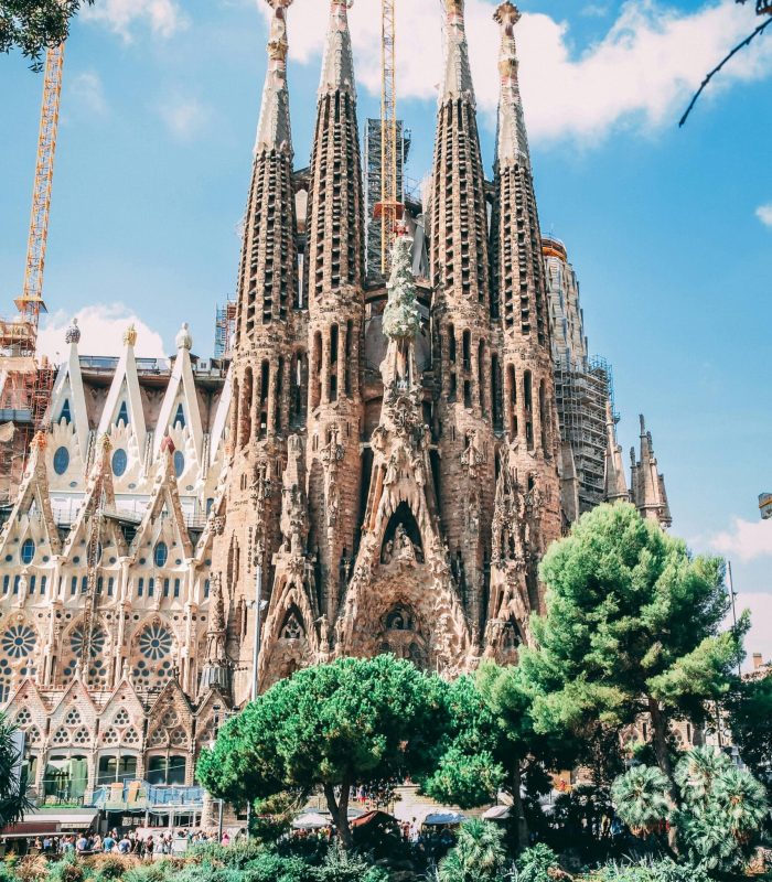 La basilique Sagrada Família à Barcelone, en Espagne, avec ses hautes tours ornées, ses façades complexes, les grues de la construction en cours, les arbres et l'étang au premier plan sous un ciel bleu lumineux.