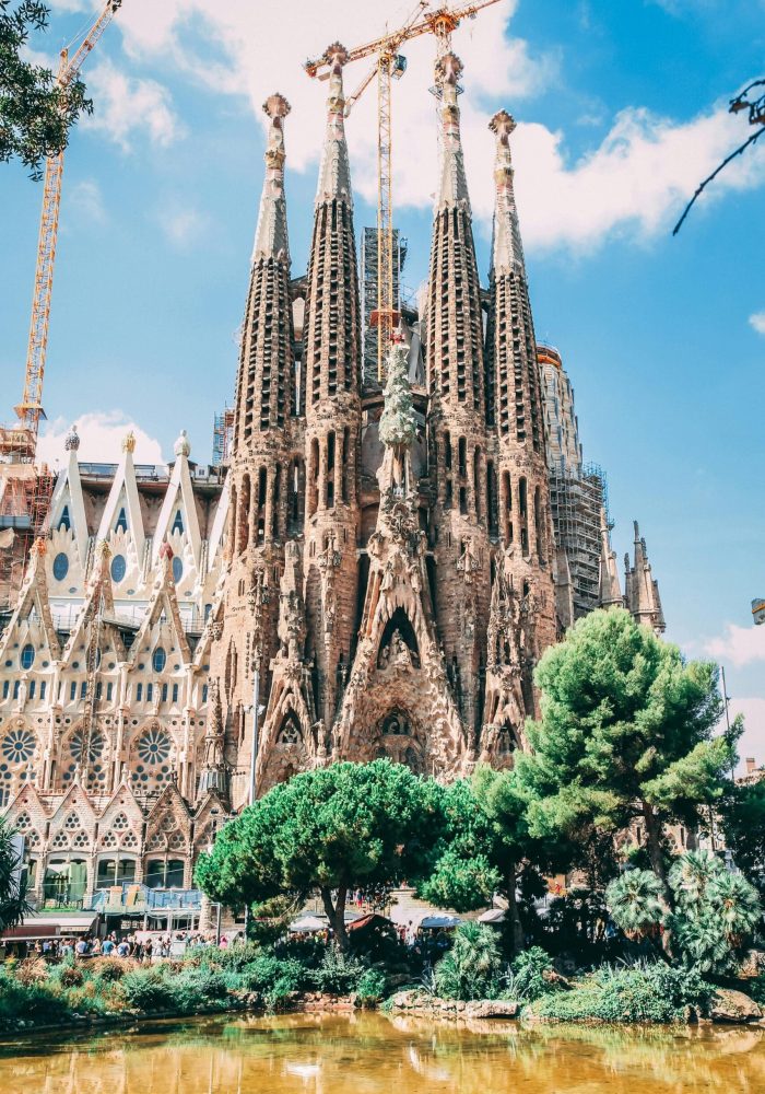 La basilique Sagrada Família à Barcelone, en Espagne, avec ses hautes tours ornées, ses façades complexes, les grues de la construction en cours, les arbres et l'étang au premier plan sous un ciel bleu lumineux.