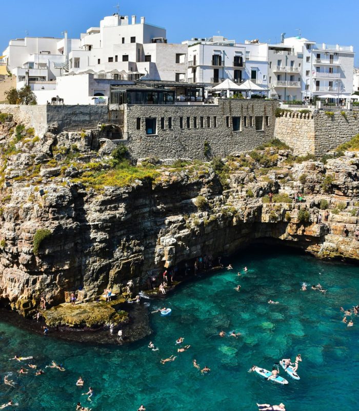 Les nageurs profitent d'une eau claire et turquoise près des falaises rocheuses, avec des bâtiments blancs et des murs de pierre perchés au-dessus, sous un ciel bleu éclatant.