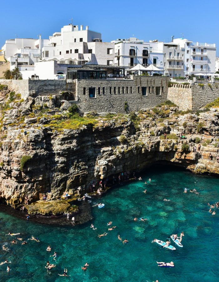 Les nageurs profitent d'une eau claire et turquoise près des falaises rocheuses, avec des bâtiments blancs et des murs de pierre perchés au-dessus, sous un ciel bleu éclatant.