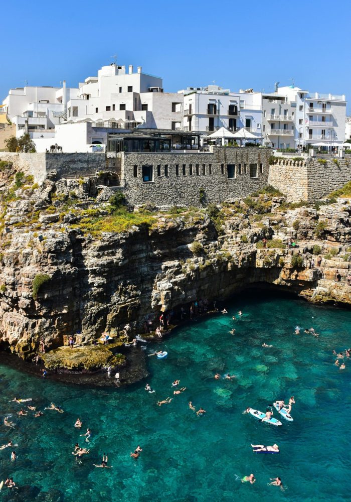 Les nageurs profitent d'une eau claire et turquoise près des falaises rocheuses, avec des bâtiments blancs et des murs de pierre perchés au-dessus, sous un ciel bleu éclatant.