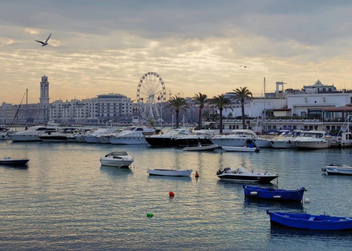 Un port avec des bateaux et des yachts amarrés, une eau calme, des palmiers le long du front de mer, une grande roue, des bâtiments urbains en arrière-plan et un ciel nuageux avec un oiseau volant au-dessus.