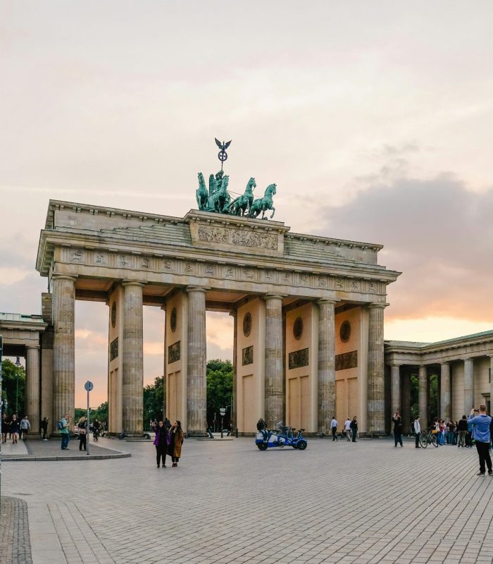 La porte de Brandebourg à Berlin au coucher du soleil, avec des groupes de personnes rassemblées autour et un ciel aux teintes orange et violettes. Le monument historique se dresse au centre d'une grande place ouverte.