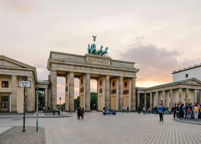 The Brandenburg Gate in Berlin at sunset, with groups of people gathered around it and a sky of orange and violet hues. The historic monument stands in the centre of a large open square.