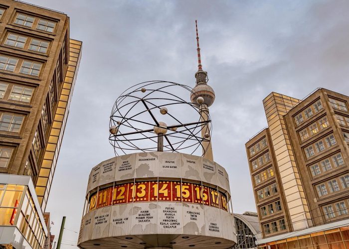 L'image montre l'horloge mondiale de Berlin sur l'Alexanderplatz avec la tour de télévision Fernsehturm en arrière-plan, entourée de bâtiments modernes sous un ciel nuageux.