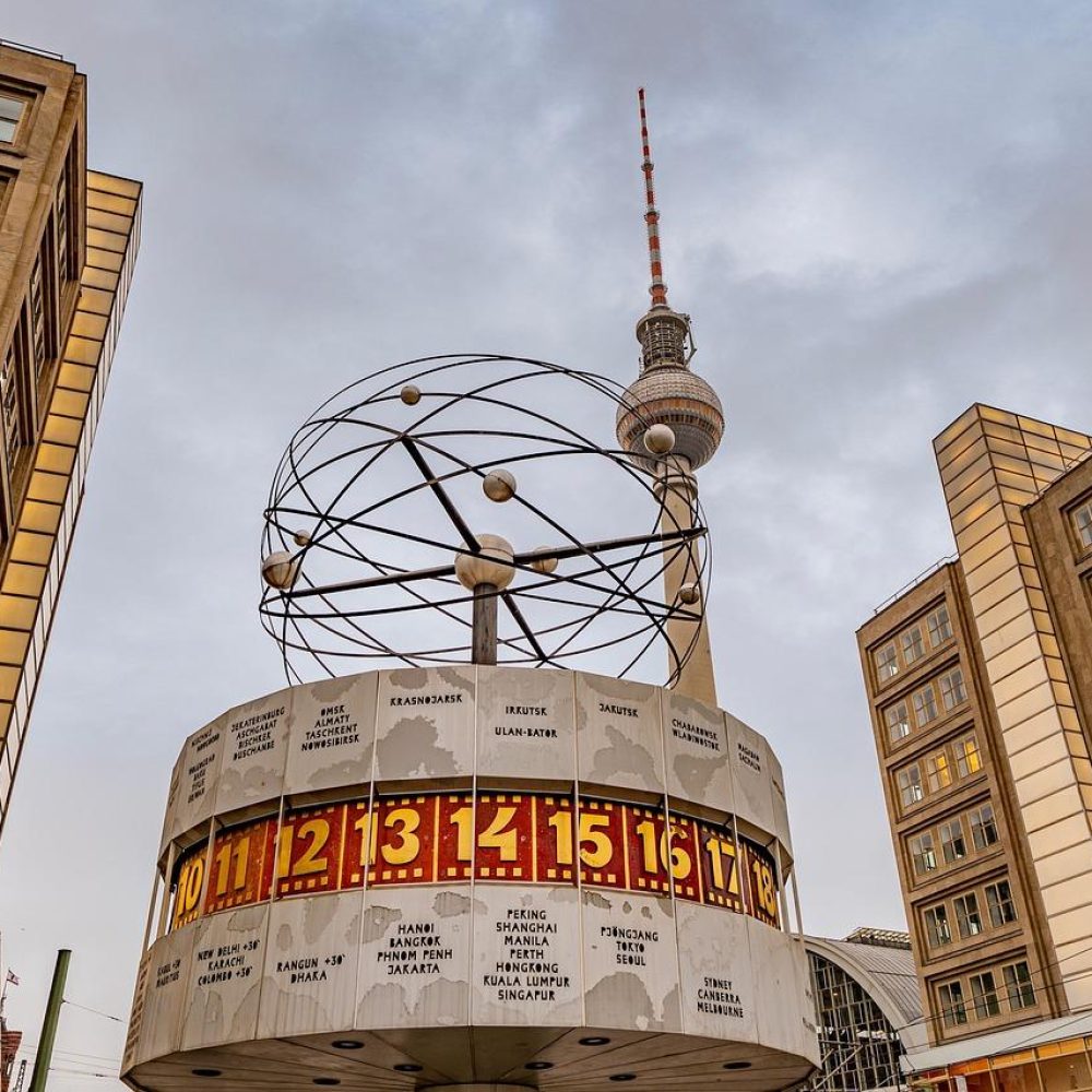 L'image montre l'horloge mondiale de Berlin sur l'Alexanderplatz avec la tour de télévision Fernsehturm en arrière-plan, entourée de bâtiments modernes sous un ciel nuageux.