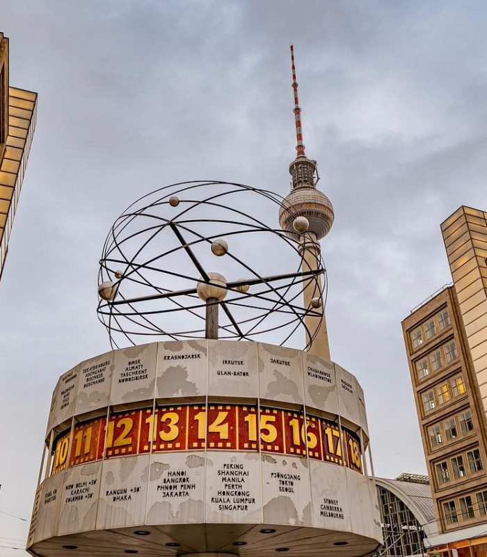 The image shows the Berlin World Clock on Alexanderplatz with the Fernsehturm television tower in the background, surrounded by modern buildings under a cloudy sky.
