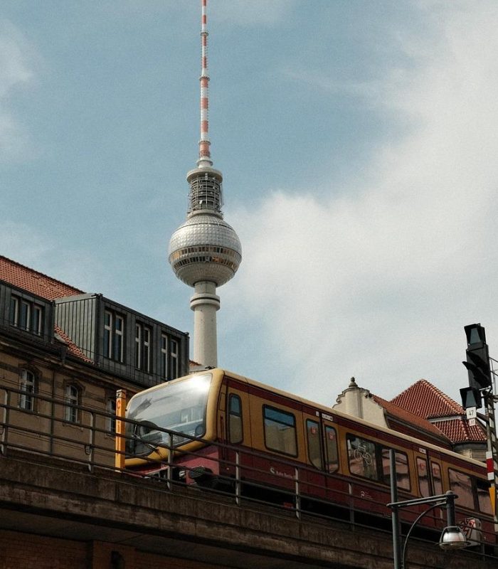 Un train jaune et rouge circule sur une voie surélevée devant la tour de télévision de Berlin, avec des bâtiments historiques et un ciel partiellement nuageux en arrière-plan.