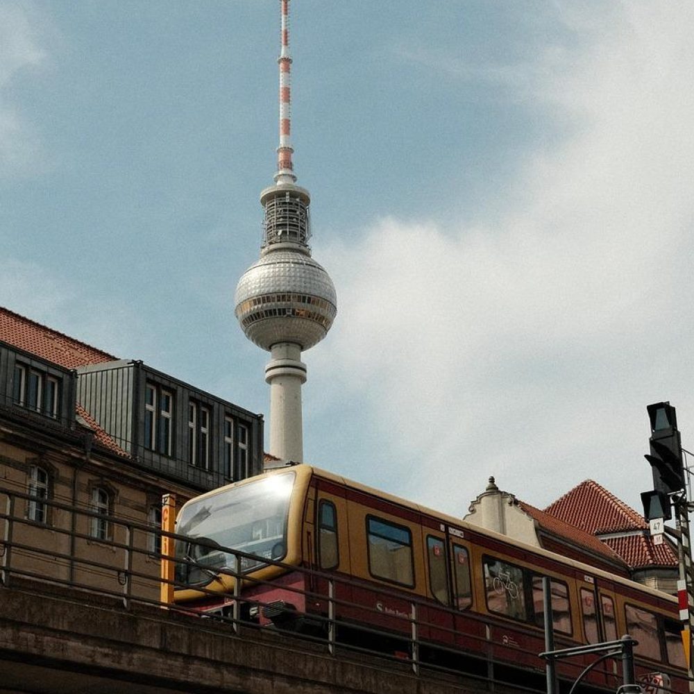 A yellow and red train runs on an elevated track in front of Berlin's television tower, with historic buildings and a partly cloudy sky in the background.