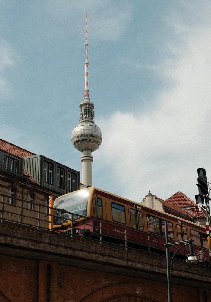 A yellow and red train runs on an elevated track in front of Berlin's television tower, with historic buildings and a partly cloudy sky in the background.