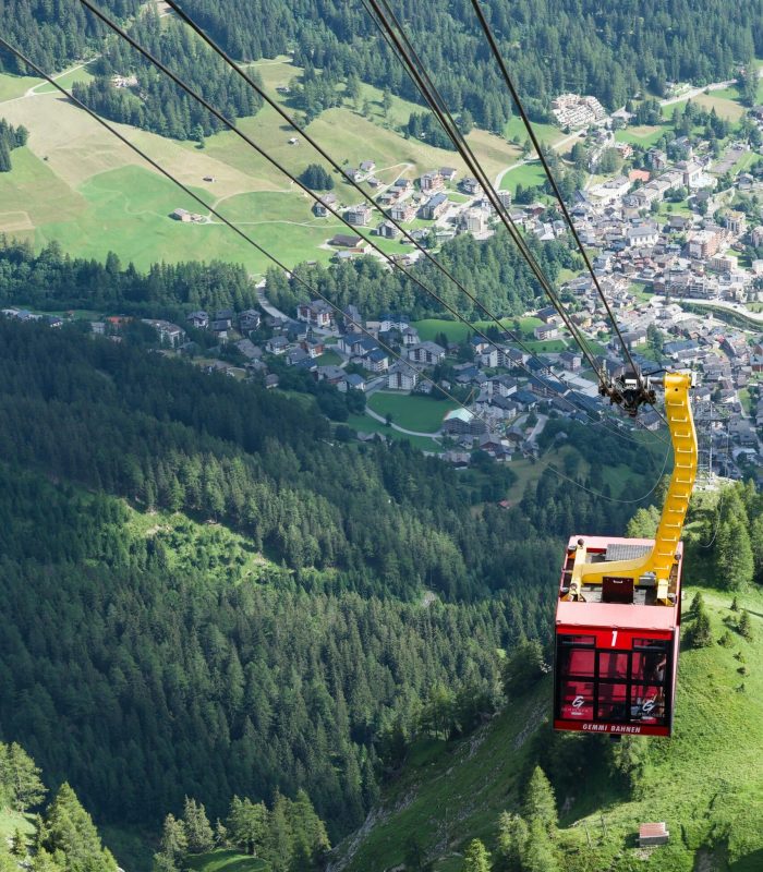 Un téléphérique rouge monte au-dessus d'une forêt verdoyante et d'une vallée, avec un village et des champs en patchwork visibles en contrebas, entourés de montagnes.
