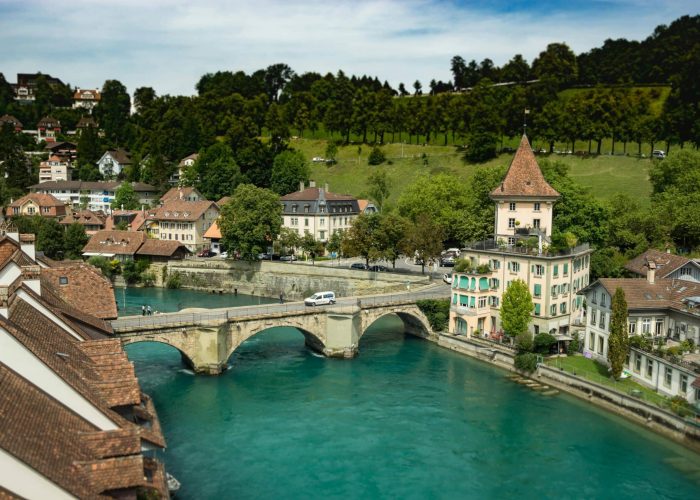 Un pont de pierre enjambe une rivière turquoise dans une ville européenne avec des bâtiments aux toits rouges, des arbres verdoyants et une tour caractéristique. La scène est tranquille et pittoresque, avec des collines en arrière-plan.