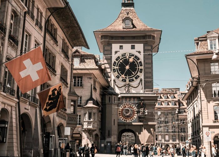 Une tour d'horloge historique se dresse au bout d'une rue pavée bordée de vieux bâtiments. Des gens marchent sous le soleil. Des drapeaux suisses et cantonaux sont accrochés à un bâtiment sur la gauche.