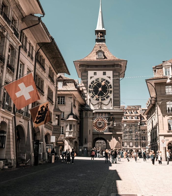 Une tour d'horloge historique se dresse au bout d'une rue pavée bordée de vieux bâtiments. Des gens marchent sous le soleil. Des drapeaux suisses et cantonaux sont accrochés à un bâtiment sur la gauche.
