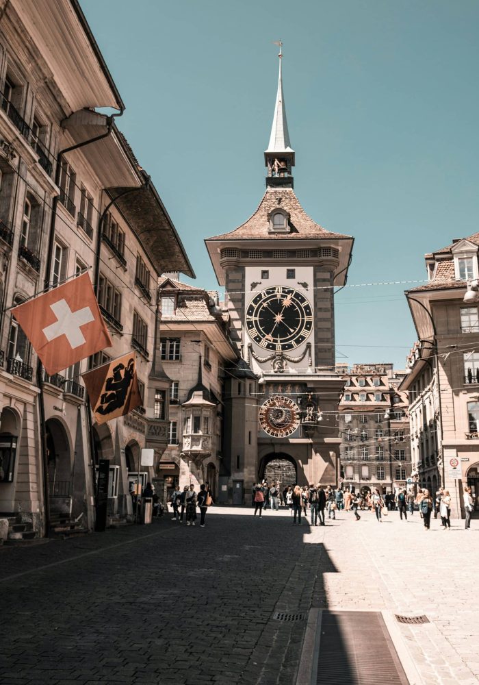 Une tour d'horloge historique se dresse au bout d'une rue pavée bordée de vieux bâtiments. Des gens marchent sous le soleil. Des drapeaux suisses et cantonaux sont accrochés à un bâtiment sur la gauche.