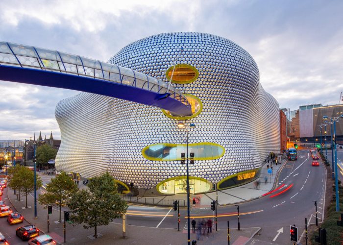 Un bâtiment futuriste à la façade incurvée, argentée, à motifs circulaires et aux fenêtres ovales se dresse le long d'une rue animée, avec une passerelle bleue pour piétons qui s'étend à son niveau supérieur. Les voitures et les lumières de la ville remplissent la scène au crépuscule.