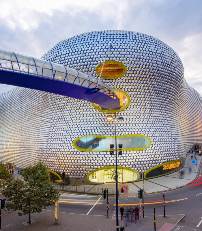 Un bâtiment futuriste à la façade incurvée, argentée, à motifs circulaires et aux fenêtres ovales se dresse le long d'une rue animée, avec une passerelle bleue pour piétons qui s'étend à son niveau supérieur. Les voitures et les lumières de la ville remplissent la scène au crépuscule.