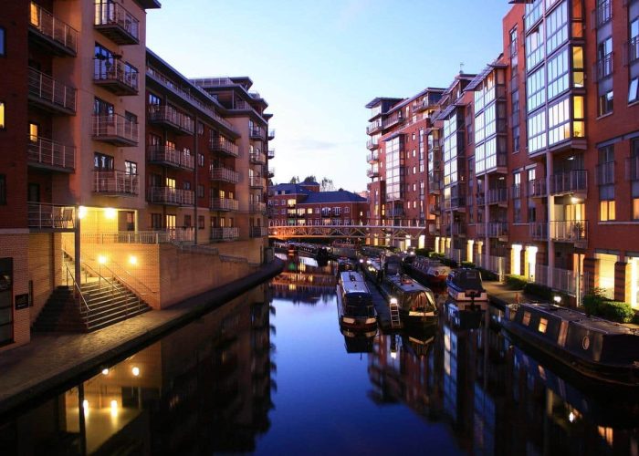 Un canal bordé d'immeubles modernes au crépuscule, avec des bateaux étroits amarrés le long de l'eau et des lumières chaudes se reflétant sur la surface calme. Au loin, une passerelle piétonne traverse le canal.