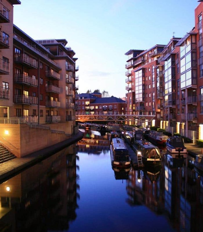 Un canal bordé d'immeubles modernes au crépuscule, avec des bateaux étroits amarrés le long de l'eau et des lumières chaudes se reflétant sur la surface calme. Au loin, une passerelle piétonne traverse le canal.