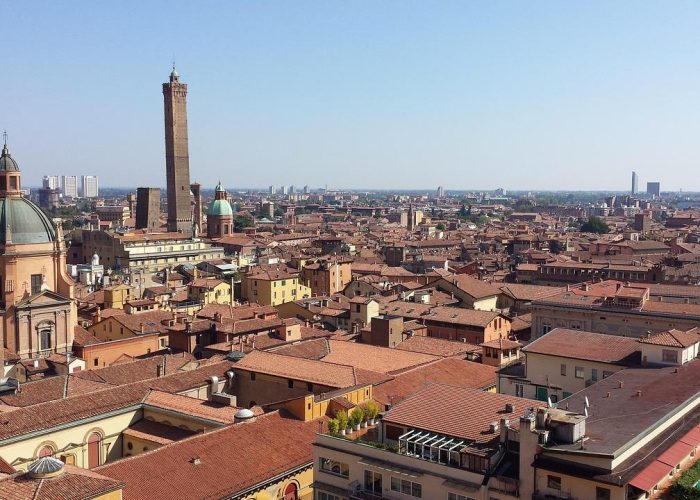 Vue panoramique de Bologne, en Italie, montrant des toits en terre cuite, deux hautes tours médiévales, des églises avec des dômes et des bâtiments de la ville sous un ciel bleu et clair.