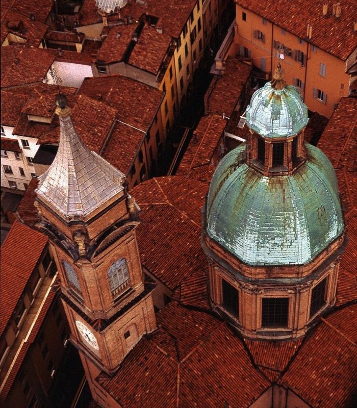 Vue aérienne d'une ville européenne historique aux toits de tuiles rouges, avec une église à dôme au toit patiné vert et une tour d'horloge adjacente parmi les bâtiments anciens environnants.