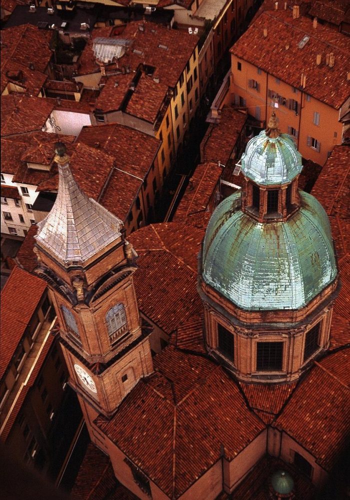 Vue aérienne d'une ville européenne historique aux toits de tuiles rouges, avec une église à dôme au toit patiné vert et une tour d'horloge adjacente parmi les bâtiments anciens environnants.